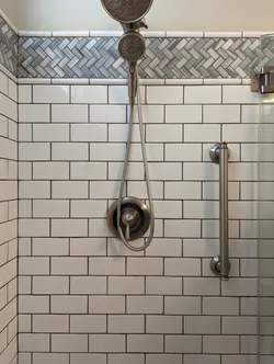 Finished tile shower enclosure with brushed nickel pressure-balance valve trim, dual rain-head and handheld showerhead combo, wall-mounted grab bar, subway tile field with herringbone marble accent border, and frameless glass door — no visible supply rough-in or failure modes, all trim finish work.