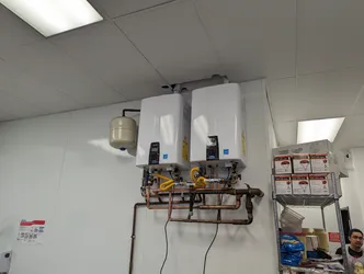 Dual tankless water heaters wall-mounted in a commercial utility space, plumbed with copper supply and return manifolds, yellow CSST gas flex connectors, ball valves throughout, and a single expansion tank on the left unit.
