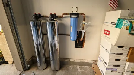 Dual-tank water softener with stainless brine/resin tanks plumbed in copper at the point of entry in a residential garage, with a blue whole-house sediment pre-filter canister and bypass valve assembly mounted to the wall.