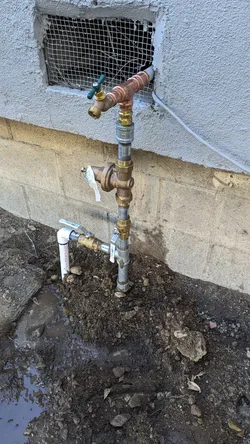 Exterior foundation wall showing a galvanized riser transitioning to copper with brass ball valves and a hose bib above grade, tying into a PVC stub-out at the base near a wet, excavated trench — galvanic corrosion risk at every dissimilar-metal union, with signs of active or recent ground saturation.
