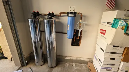Dual-tank water softener system with a whole-house sediment pre-filter canister plumbed in copper at the main service entry in a garage, bypass valves installed, no visible corrosion or scale on exposed fittings.
