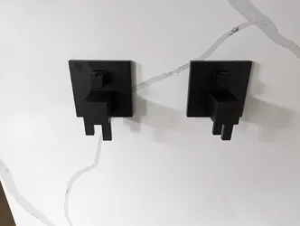 Two matte black square-handle in-wall valve trim kits mounted on a porcelain tile surround, supply valves recessed into wall with concealed rough-in bodies, no visible corrosion or material failures, protective tape still on tile edges indicating recent finish install.