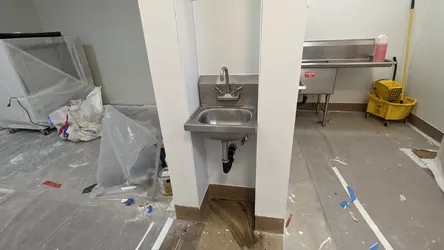 Commercial stainless hand sink with deck-mount two-handle faucet and black ABS P-trap rough-in during an active interior remodel, drain not yet connected to floor, construction debris visible throughout.