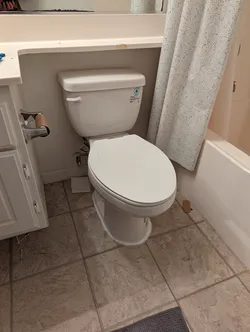 Two-piece elongated toilet with braided stainless supply line set in a finished bathroom, toilet not caulked to tile floor, wax ring seal condition unknown, adjacent tub/shower combo with curtain rod visible.