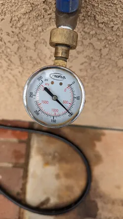 ProFlo brass hose-bib pressure gauge threaded onto an exterior hose bibb against a stucco wall, reading approximately 100 psi static supply pressure, with light surface oxidation on the brass fitting.