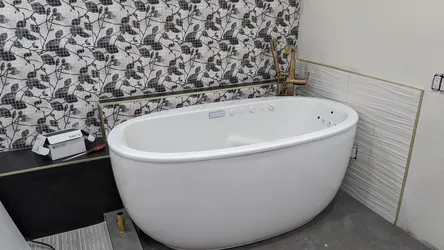 Freestanding acrylic jetted soaker tub set in finish-stage bathroom with brushed-gold floor-mount filler rough-in stubbed up, large-format tile and mosaic accent wall partially installed, no visible supply connections finalized yet.