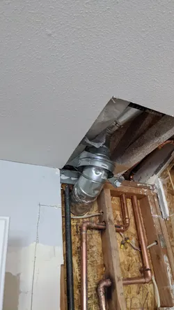 Opened wall and ceiling in a residential interior showing copper supply lines with soldered elbows and tees roughed into OSB-sheathed framing, alongside a black ABS or gas line and a double-wall metal B-vent or dryer duct terminating through the ceiling.