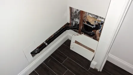 Wall opened in a finished bathroom corner revealing ABS or black PVC drain line running along the base of the wall with a 90-degree sweep fitting, alongside exposed fiberglass batt insulation, foil vapor barrier, and what appears to be white PVC supply lines with push-fit fittings visible in the upper wall cavity — indicative of a leak investigation or drain reroute.