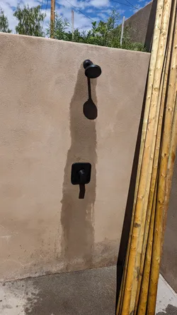 Outdoor shower rough-in on a stucco-finished masonry wall with matte black trim-kit valve and fixed showerhead, supply lines concealed in wall, no visible corrosion, adjacent bamboo privacy screen, appears to be a completed exterior installation.