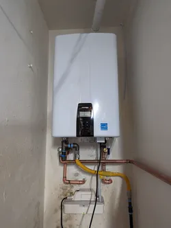 Navien tankless water heater wall-mounted in a utility alcove, plumbed with copper supply lines and a corrugated stainless gas flex, blue SharkBite push-fit fittings on the cold inlet, PVC condensate drain exiting the top, no visible corrosion but wall shows prior moisture staining at the base.