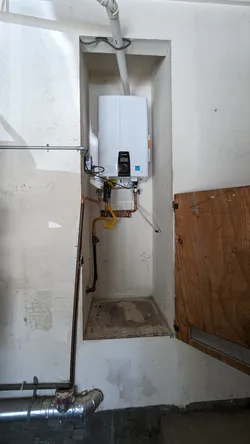 Tankless gas water heater mounted in a recessed drywall alcove with copper water supply lines, yellow CSST flexible gas connector, white PVC combustion air/exhaust venting overhead, and galvanized steel drain piping at the base.
