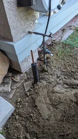 Exterior slab excavation showing a copper stub-out with foam pipe insulation rising from grade alongside an existing hose bib fed by galvanized supply line showing surface corrosion at the fitting connections.