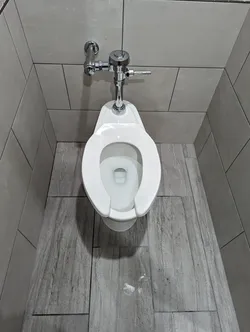 Wall-hung commercial toilet with chrome manual flushometer valve on a chrome supply stub-out, tile-finished commercial restroom installation, no visible corrosion or scale, seat missing, minor water pooling on the floor at the base.