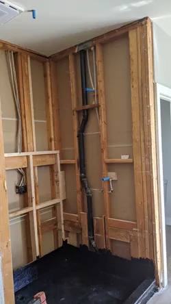 Bathroom rough-in remodel with drywall removed, showing ABS black drain stack with a 90-degree sweep fitting, blue and white PEX supply lines stubbed out at a washer or utility box location, and NM electrical cable roughed in adjacent to framing.