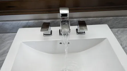 Chrome widespread lavatory faucet with square lever handles on a white undermount vitreous china sink set in marble countertop, running water, no visible corrosion or mineral scale on the spout or handles.