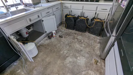 Kitchen cabinet base opened post-water-loss event showing an under-sink whole-house or point-of-use filtration canister with supply lines, flooring removed to concrete slab, and multiple air movers deployed for structural drying.